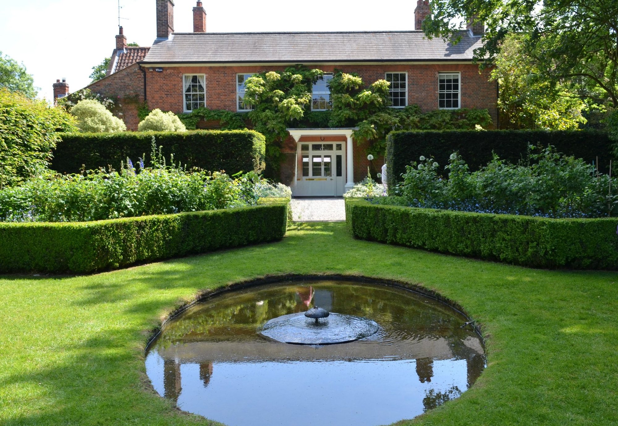 An English garden with clipped hedges, a circular pond and a wide lawn in front of a brick house